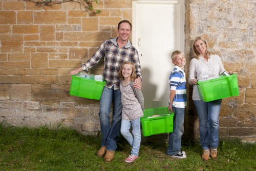 A family of four standing outside a stone building, each person holding a large, bright green plastic storage box used for rubbish or household waste collection. The father, dressed in a checkered shirt and jeans, stands on the left side with a wide smile, holding one box at his side. Next to him, a young girl with curly hair wearing a grey sweater and jeans is smiling at the camera, holding another green bin. To her right, a boy in a blue and white striped shirt and jeans stands slightly behind, also holding a green box. On the far right, the mother, dressed in a white blouse and blue jeans, holds a green waste container and looks content. The scene takes place on a grassy area adjacent to a historic stone wall with a white door visible behind the family. The overall setting appears to be a residential outdoor space, illustrating a typical rubbish collection or clearance activity, with the family ready to participate in waste disposal or clearance, fitting within the context of Knightsbridge House Clearance's rubbish removal services in London.