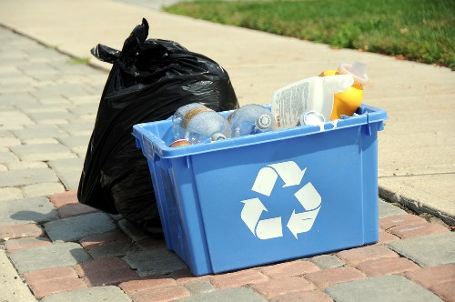 On a paved driveway in a residential area, there is a blue recycling bin with a white recycling symbol on the front, filled with empty plastic bottles and containers. Next to the bin, a black plastic trash bag is placed, slightly leaning against the bin. The plastic bottles in the bin are transparent with some showing labels, and the containers include yellow and white plastic packaging. In the background, a grassy lawn is visible, alongside a narrow sidewalk extending into the distance. The scene appears to be part of a rubbish removal collection or disposal process, reflecting typical suburban rubbish management practices in the UK. The overall lighting indicates a daytime setting with natural light illuminating the objects clearly, emphasizing the textures and colors of the rubbish. The image visually aligns with services offered by Knightsbridge House Clearance for gathering and disposing of household waste or recyclable rubbish.