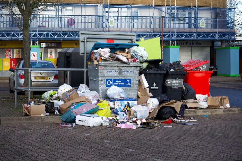 A black wheeled rubbish bin with a white label and the text 'ST. JOHN'S' printed on the front, positioned on a sidewalk at night. The bin's lid is open, revealing mixed waste including paper, cardboar